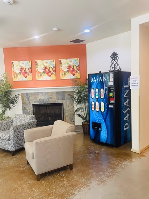 A cozy seating area with two armchairs in front of a stone fireplace, flanked by two potted plants. Above the fireplace are three colorful floral paintings on an orange accent wall. To the right, there is a blue Dasani vending machine against a white wall.
