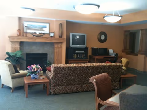 A cozy living room area with a patterned sofa, armchairs, a wooden coffee table with a flower arrangement, a fireplace with a wooden mantle, a TV on a stand, and a desk with a chair in the background. The walls are painted in warm tones and there are ceiling lights providing illumination.