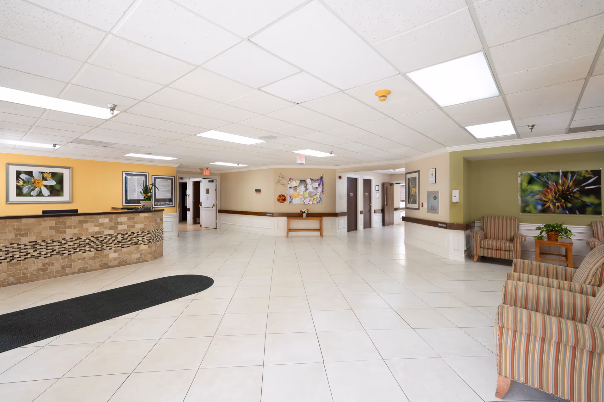 A spacious and well-lit reception area in a senior living facility with a tiled floor, a stone front desk on the left, and a seating area with striped armchairs and a small table with a plant on the right. The walls are painted in soft yellow and green tones, decorated with framed floral artwork. There is a bulletin board on the far wall and several doorways leading to other parts of the facility.