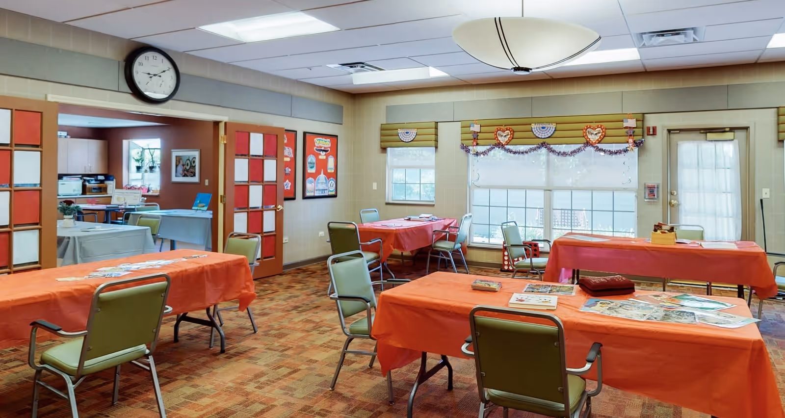 A bright room with several tables covered in orange tablecloths and green chairs arranged around them. The room has large windows with blinds and decorative heart-shaped garlands. A clock is mounted on the wall above a set of double doors leading to another room with more tables and chairs. The floor is carpeted with a patterned design, and the ceiling has recessed lighting and a large round light fixture.