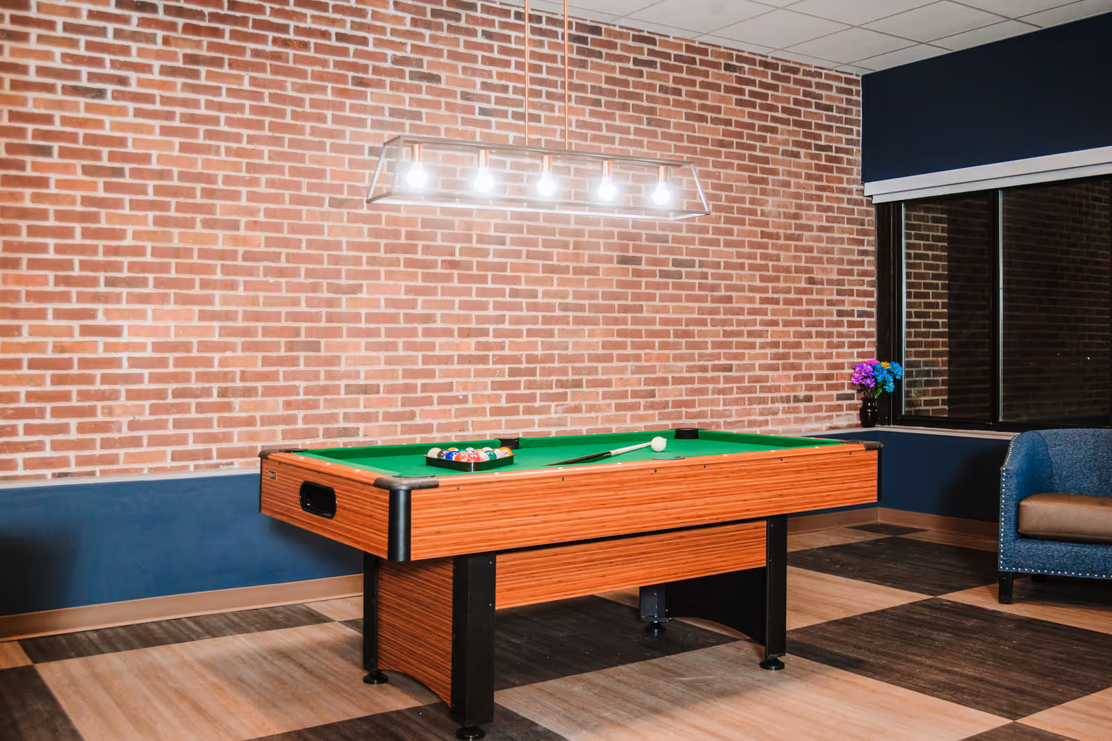A recreational room with a pool table in the center, a brick wall in the background, a modern hanging light fixture above the table, a window with a dark blue valance, a small vase with colorful flowers on the windowsill, and a blue upholstered chair with a brown cushion to the right.
