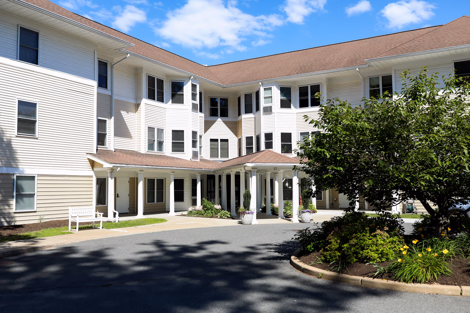 Exterior view of a three-story white retirement community building with a covered entrance supported by white columns. There is a circular driveway with landscaped greenery and a tree in front of the building under a blue sky with some clouds.