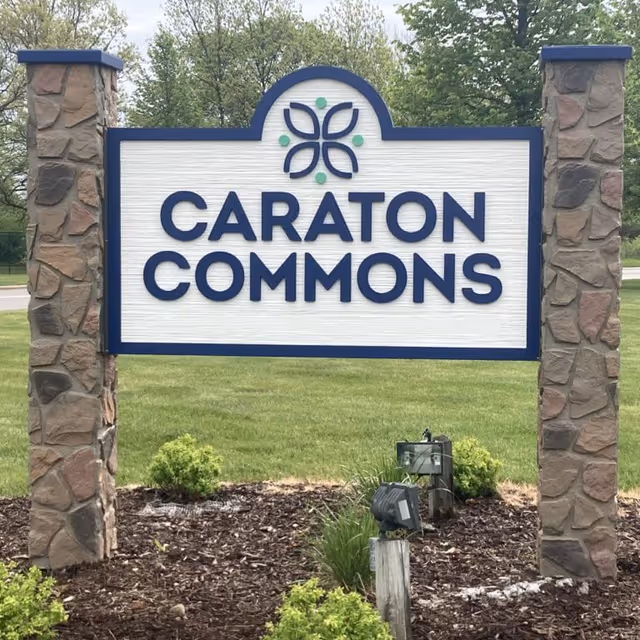 Outdoor sign for Caraton Commons mounted between two stone pillars with grass and small shrubs around the base, and trees and a road in the background.