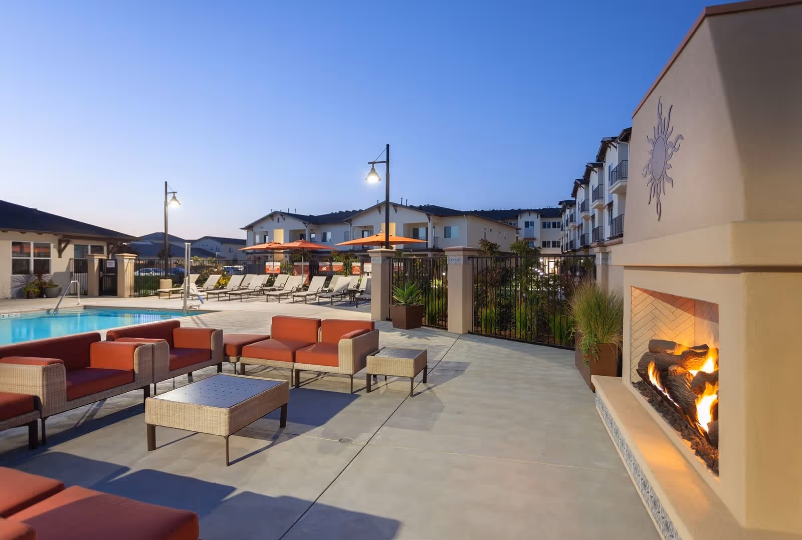 Outdoor poolside seating area with red-cushioned chairs, a lit fireplace, and an apartment building in the background at dusk.