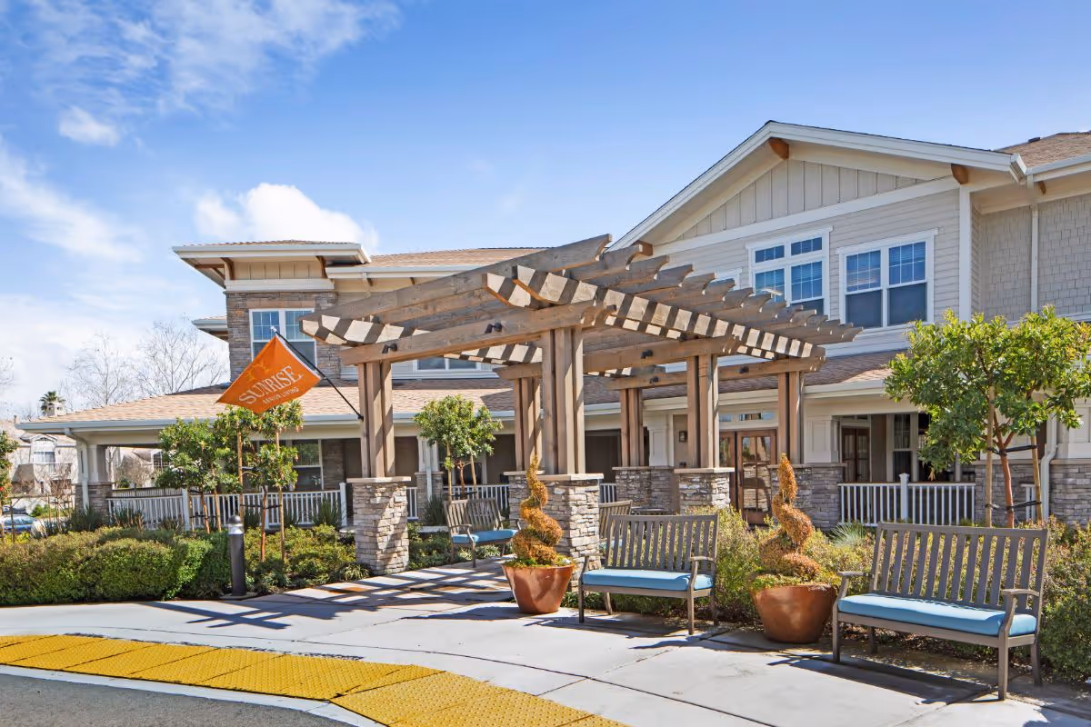 Exterior view of Ivy Park at Pleasanton senior living facility featuring a wooden pergola entrance with stone pillars, benches with blue cushions, potted plants, and a clear blue sky.