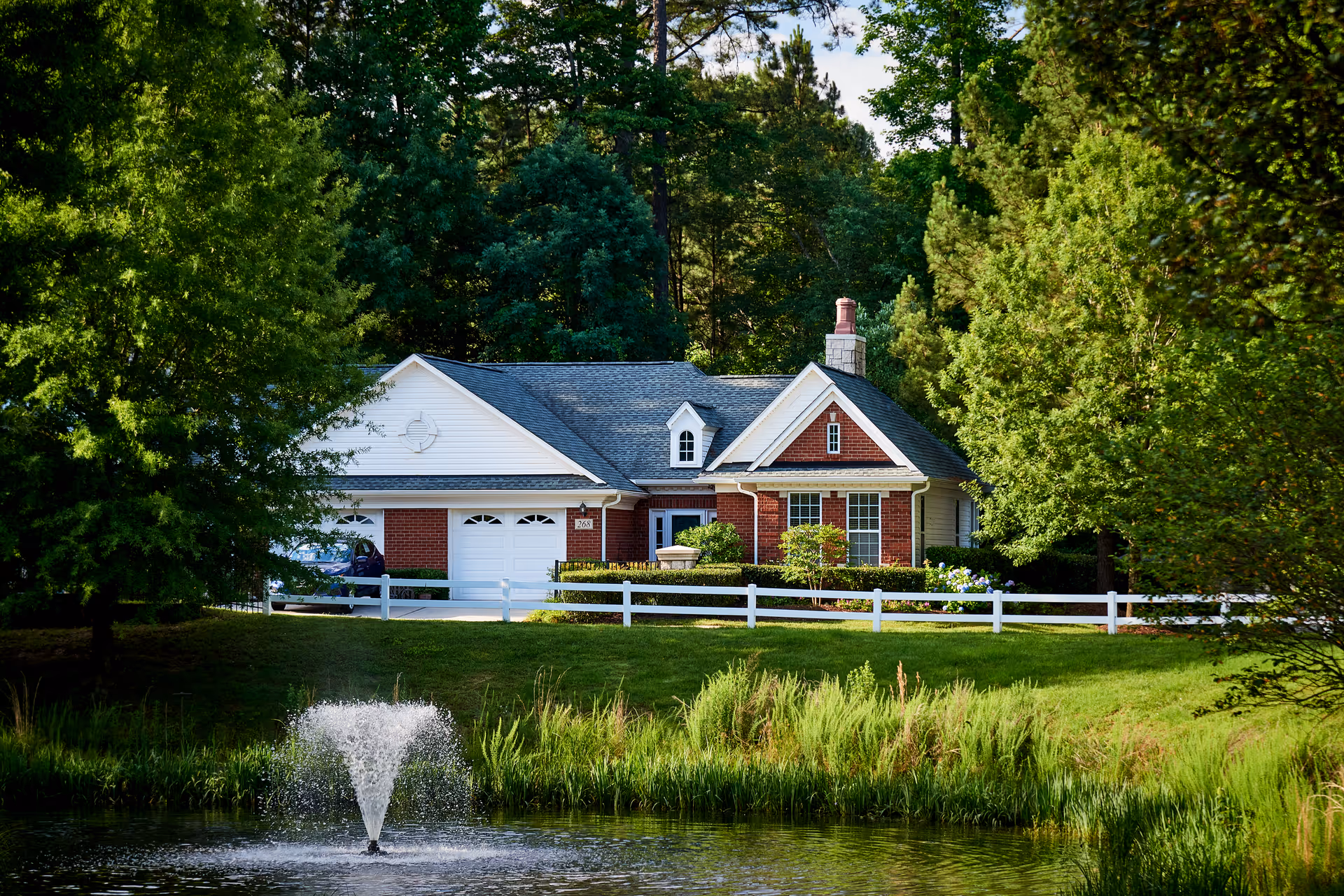 A single-story brick and white siding house surrounded by trees and greenery with a white fence in front. In the foreground, there is a pond with a water fountain spraying water upwards.