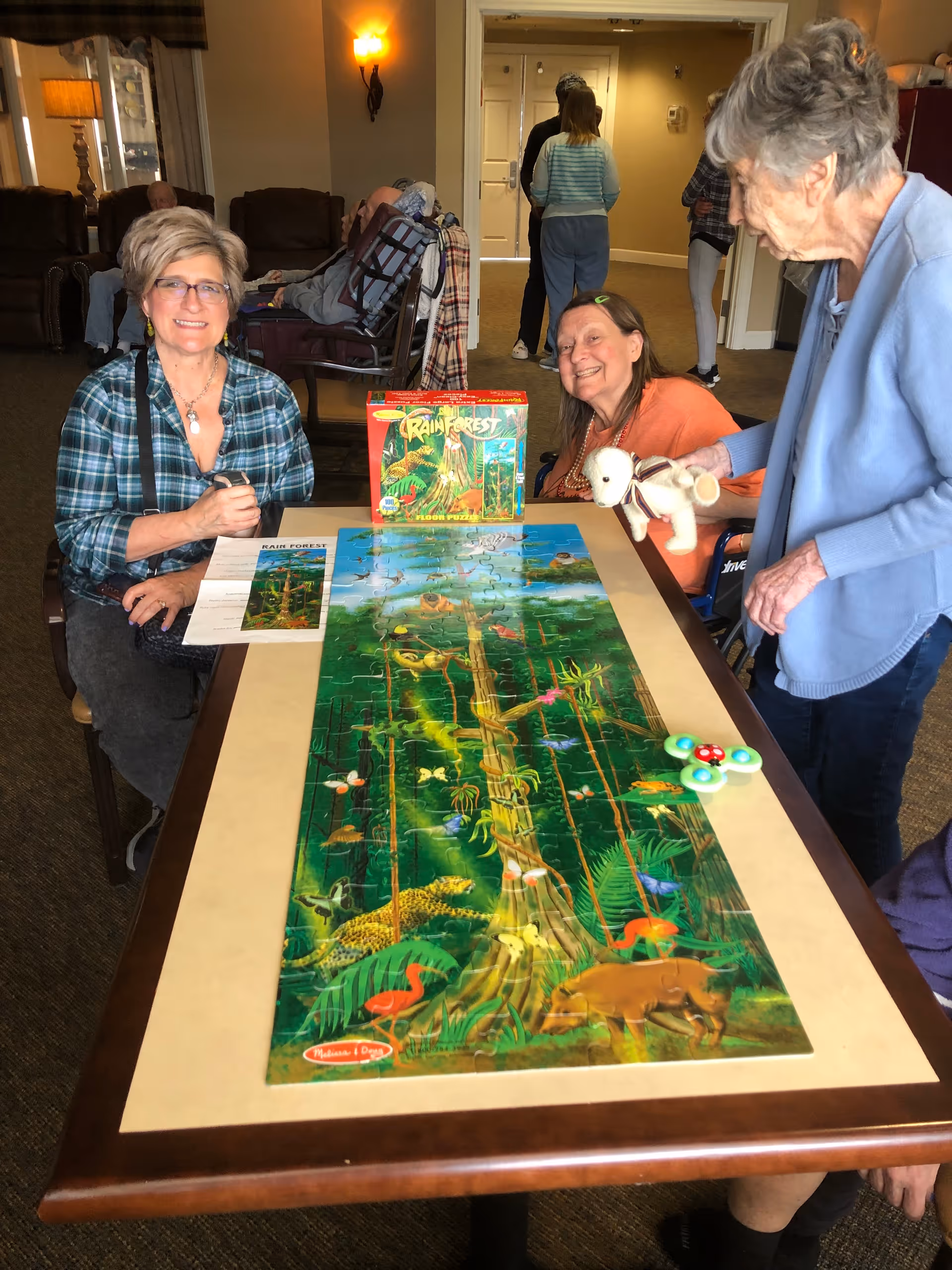 Three older adults gathered around a table working on a large rainforest jigsaw puzzle in a communal living room.