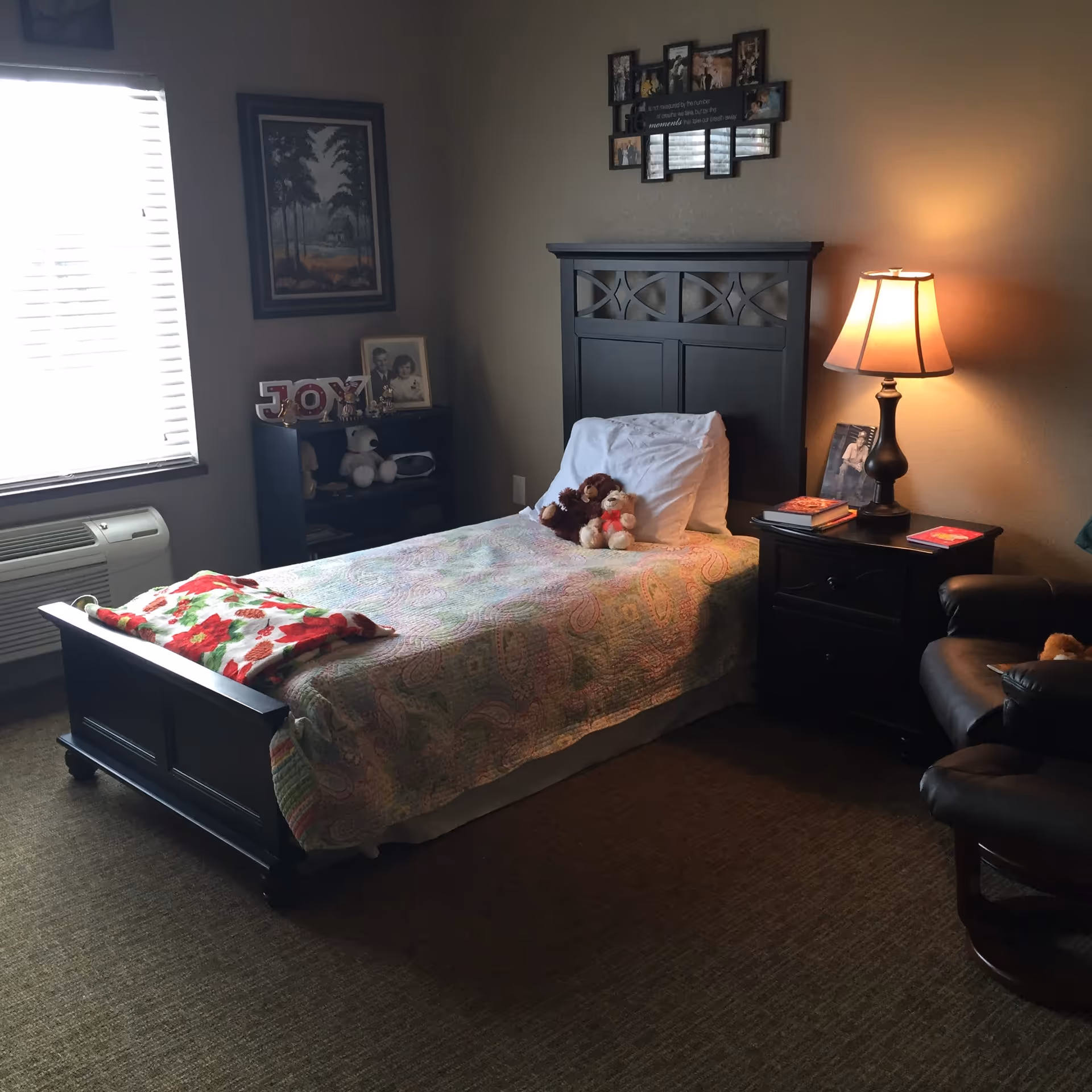 A cozy bedroom with a single bed covered in a pastel-colored quilt and a red and green blanket folded at the foot. Two teddy bears rest on the pillow. To the right of the bed is a dark wooden nightstand with a lamp, books, and a framed photo. A comfortable dark leather chair is partially visible. On the left side, a window with blinds lets in natural light, and below it is an air conditioning unit. A small dark wooden shelf holds decorative items including a 'JOY' sign and framed photos. The walls are painted beige and decorated with framed pictures.