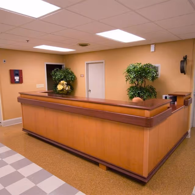 Reception desk area in a senior living facility with a wooden counter, two potted plants, beige walls, and a tiled floor under fluorescent ceiling lights.