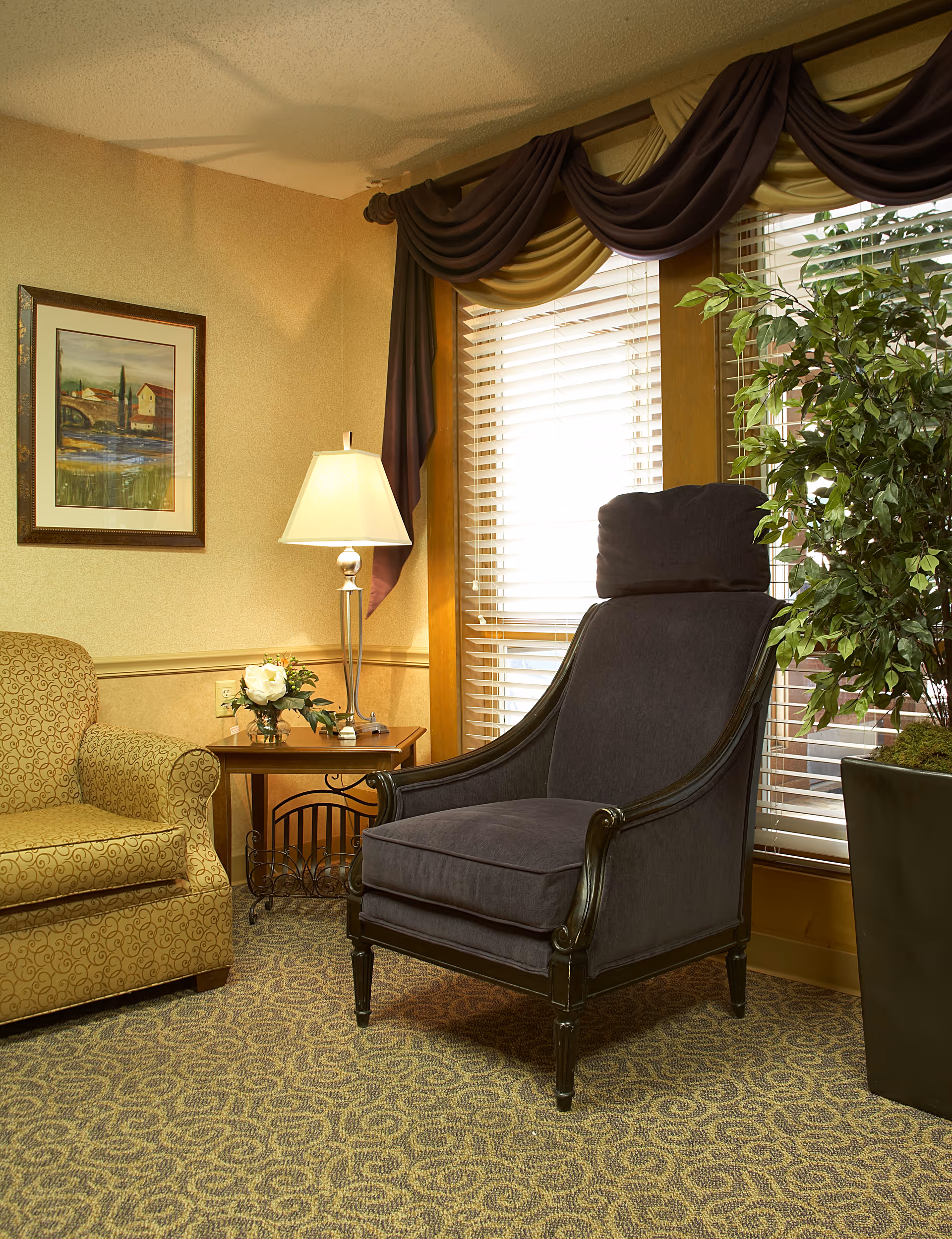 A cozy living room area with a patterned beige armchair, a dark upholstered armchair with a headrest, a wooden side table with a lamp and a small flower arrangement, a large potted plant, and a window with blinds and decorative drapes.