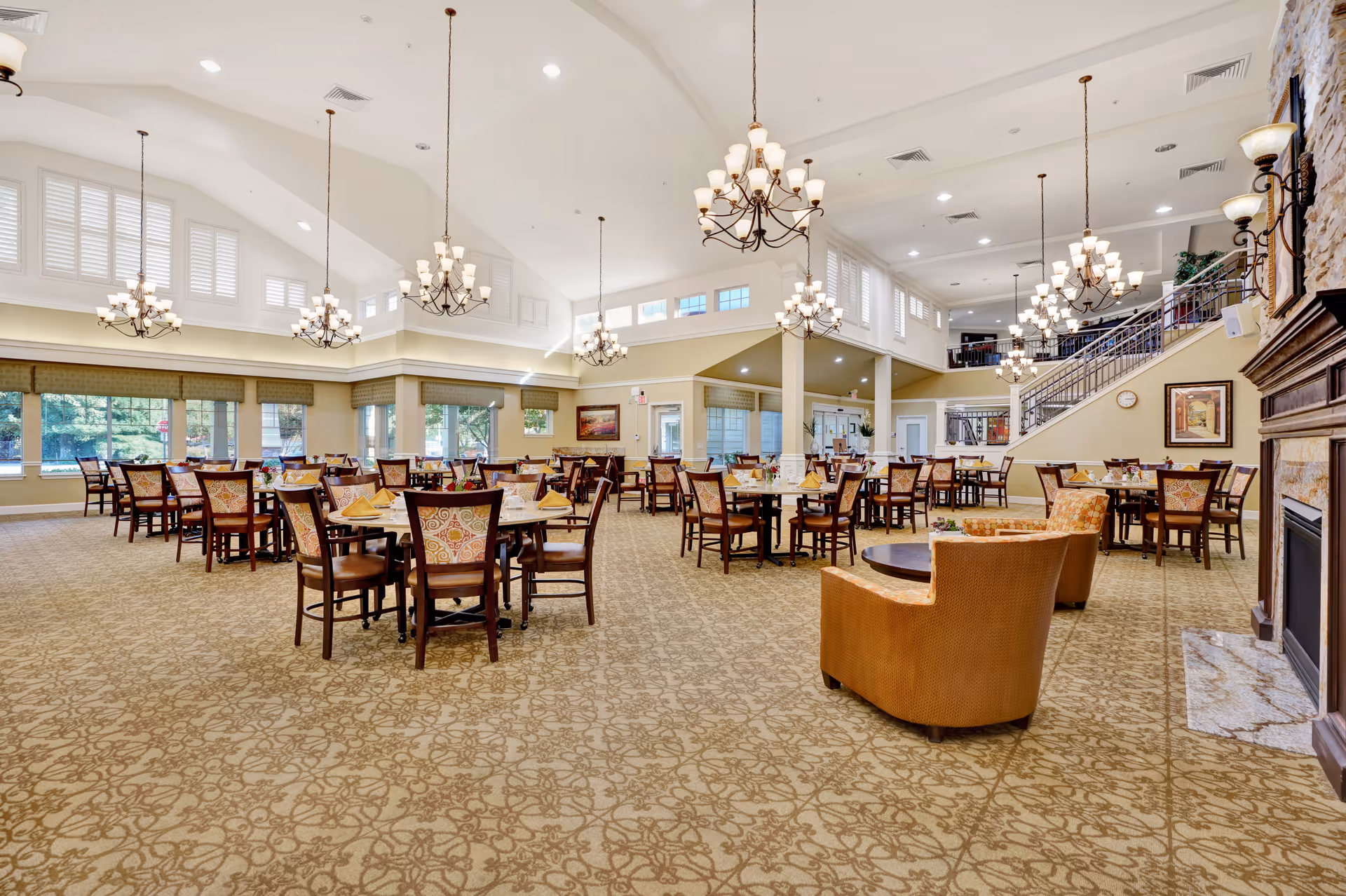 Spacious dining room with multiple round tables and chairs arranged neatly. The room features large windows with natural light, high vaulted ceilings with multiple chandeliers, patterned carpet, and a fireplace on the right side. There are also two upholstered chairs and a small round table near the fireplace, and a staircase leading to an upper level in the background.