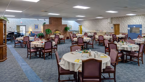 Spacious dining room with round tables set for meals, chairs, and a central brick fireplace in a senior facility.
