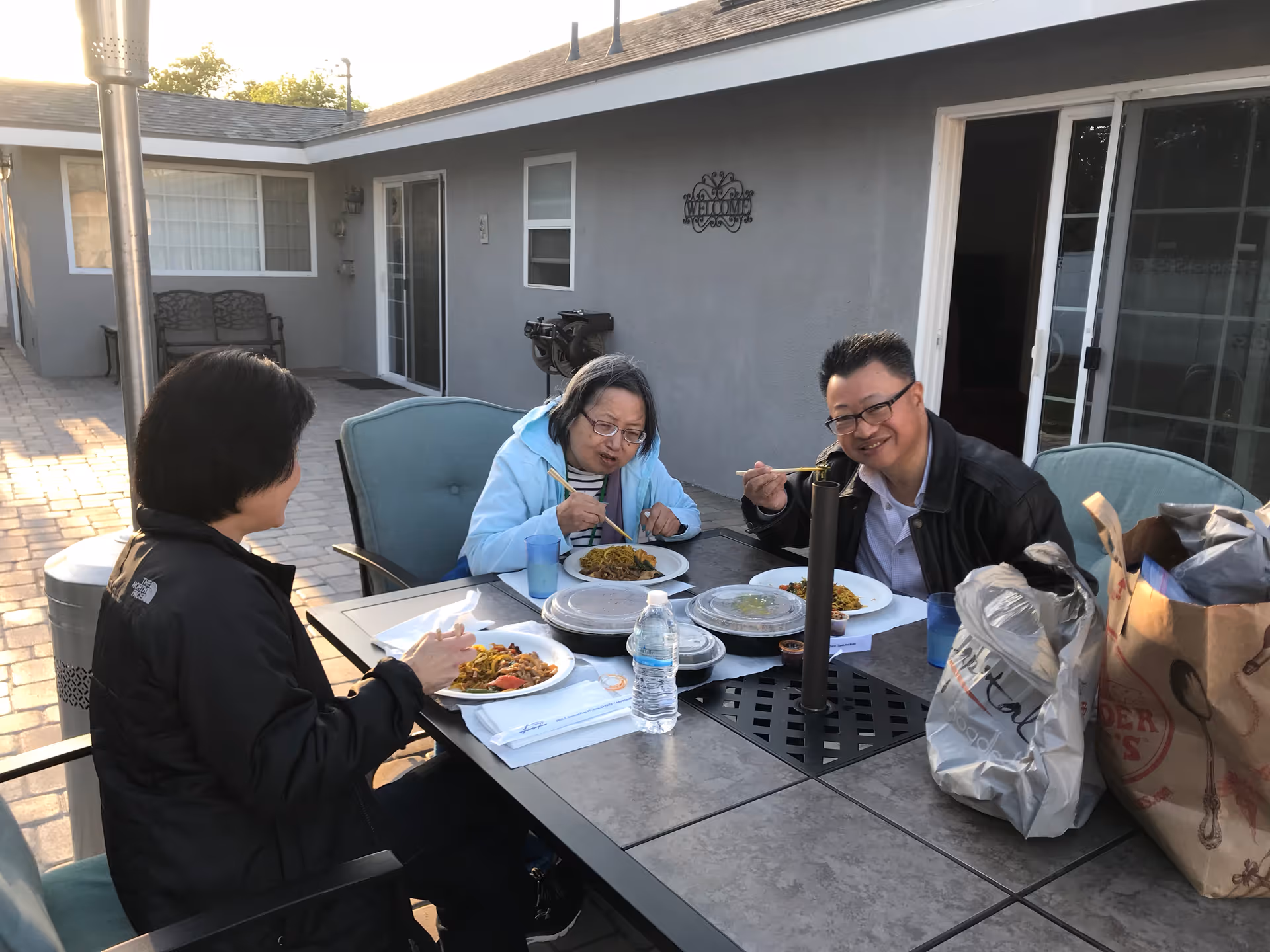 Three people sitting around an outdoor table enjoying a meal together. The table has plates of food, water bottles, and some shopping bags. They are seated on a patio area next to a gray building with sliding glass doors and windows.