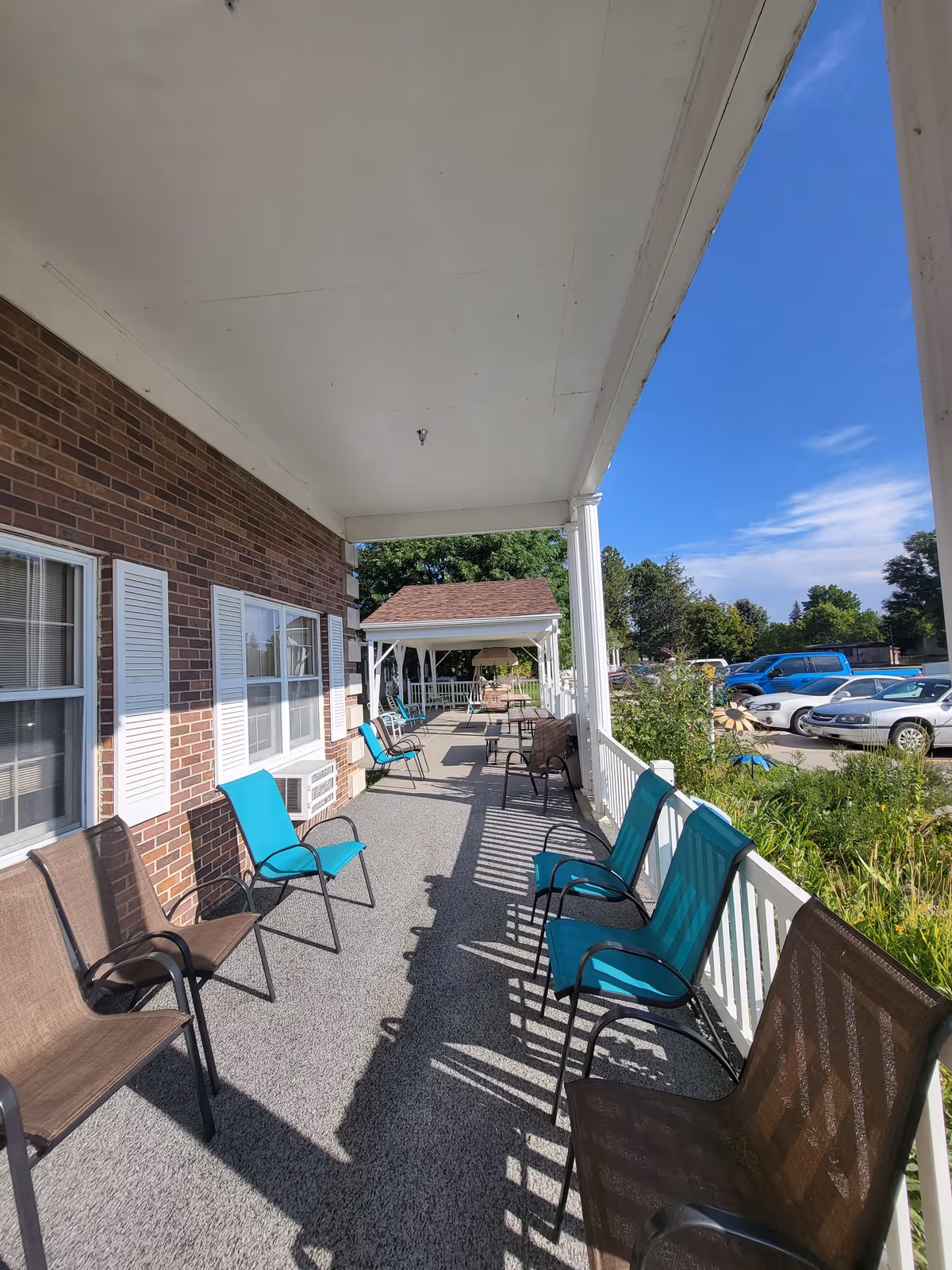 A covered outdoor porch area at Ponderosa Villa with multiple chairs arranged along the railing and brick wall. The porch overlooks a parking lot with several cars and some greenery. The sky is clear and blue.