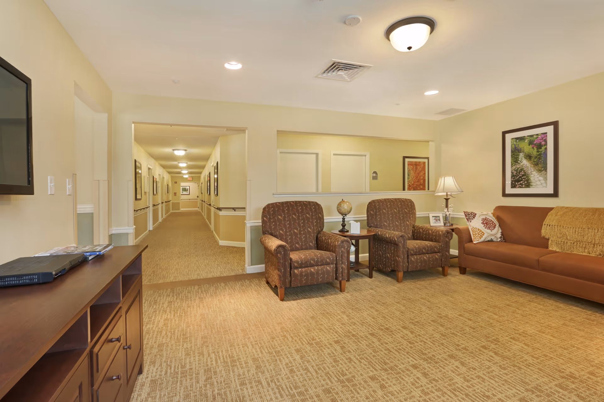 A well-lit senior living facility common area with two patterned armchairs and a brown sofa arranged around a small wooden side table with a lamp and decorative items. The room has beige walls and carpet, a framed picture on the wall, and a hallway leading to other rooms in the background.