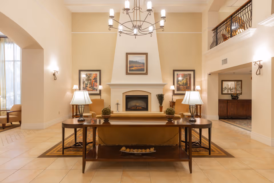 A spacious and warmly lit living room area in Casa de las Campanas featuring a central fireplace with a framed painting above it. The room has beige walls and tiled floors, with two armchairs and side tables with lamps on either side of the fireplace. A wooden console table with decorative items and two lamps is positioned behind a sofa facing the fireplace. There is a chandelier hanging from the ceiling and a balcony railing visible on the upper right side.