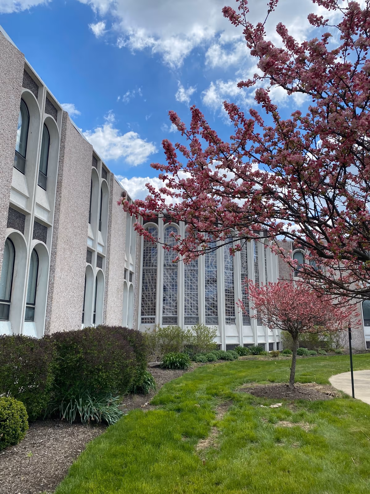 Exterior view of a senior living facility building with large arched windows and a well-maintained garden featuring green grass, bushes, and blooming pink trees under a partly cloudy blue sky.