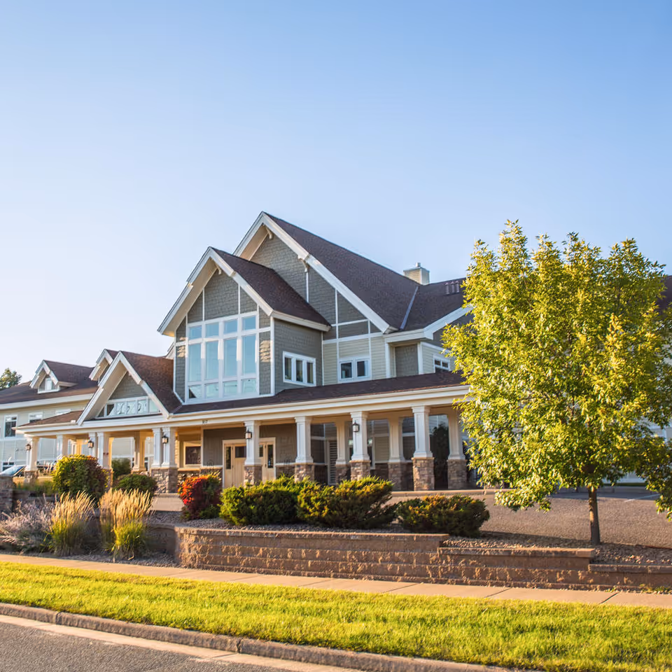 Exterior view of a large, modern assisted living facility building with multiple peaked roofs, large windows, a covered entrance supported by columns, and well-maintained landscaping including bushes, ornamental grasses, and a tree. The sky is clear and blue.