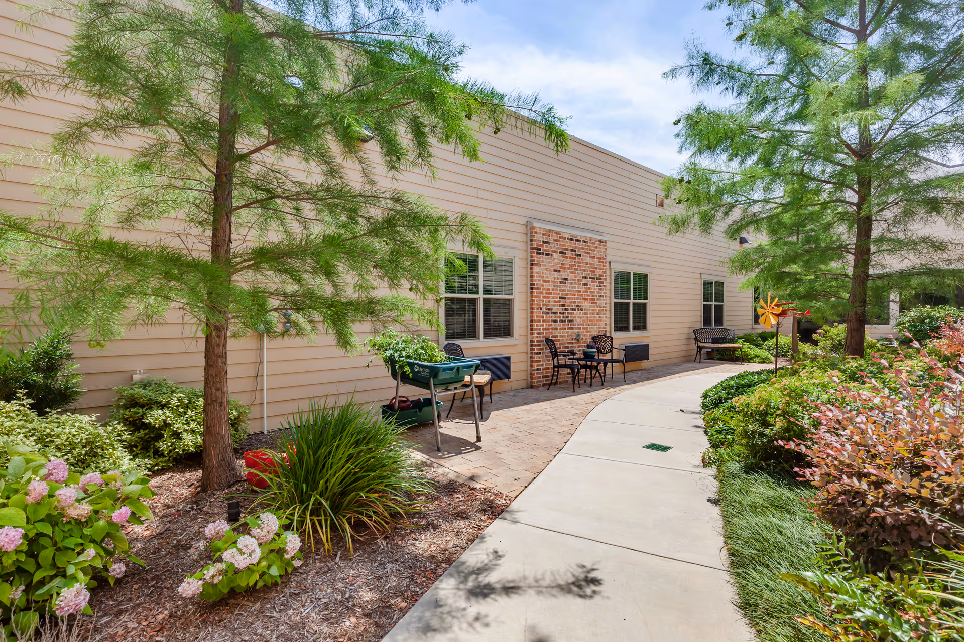 Outdoor walkway beside a beige building with windows and brick accents, surrounded by landscaped greenery including trees, bushes, and flowering plants. There are several seating areas with chairs and tables along the walkway under a partly cloudy sky.