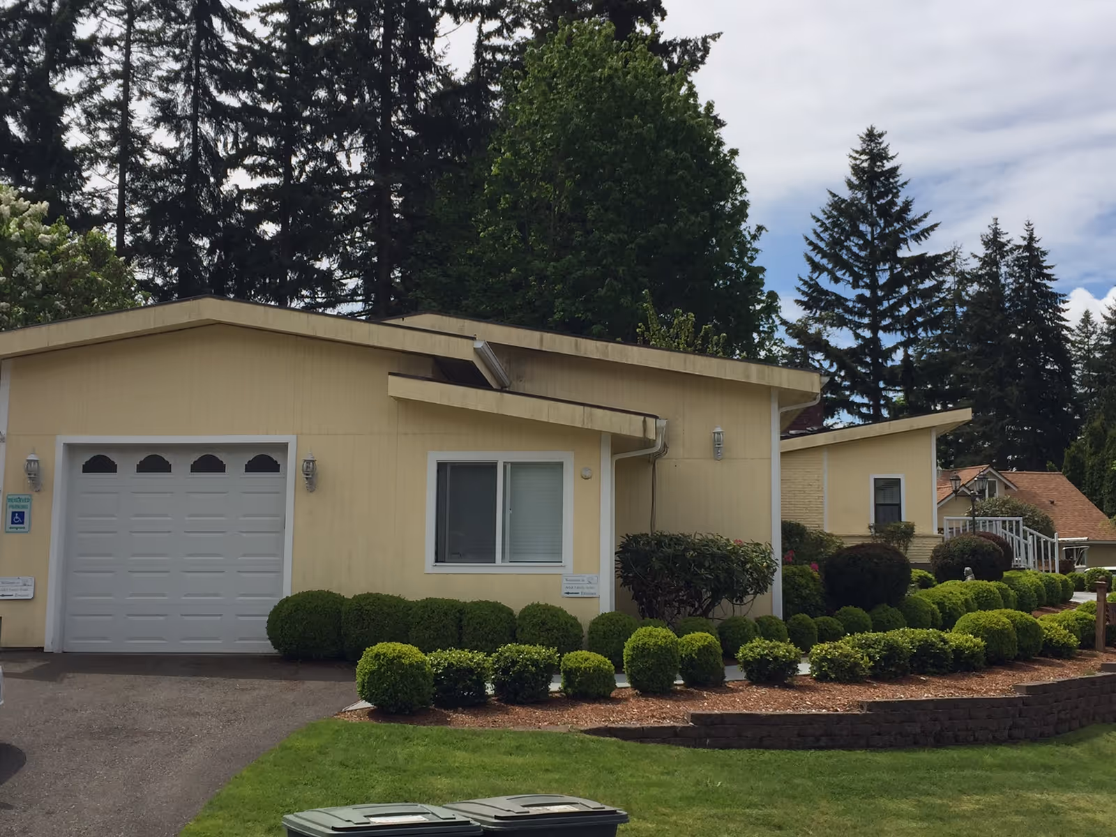 Front exterior of a single-story pale yellow house with an attached garage, trimmed shrubs, and tall trees in the background.