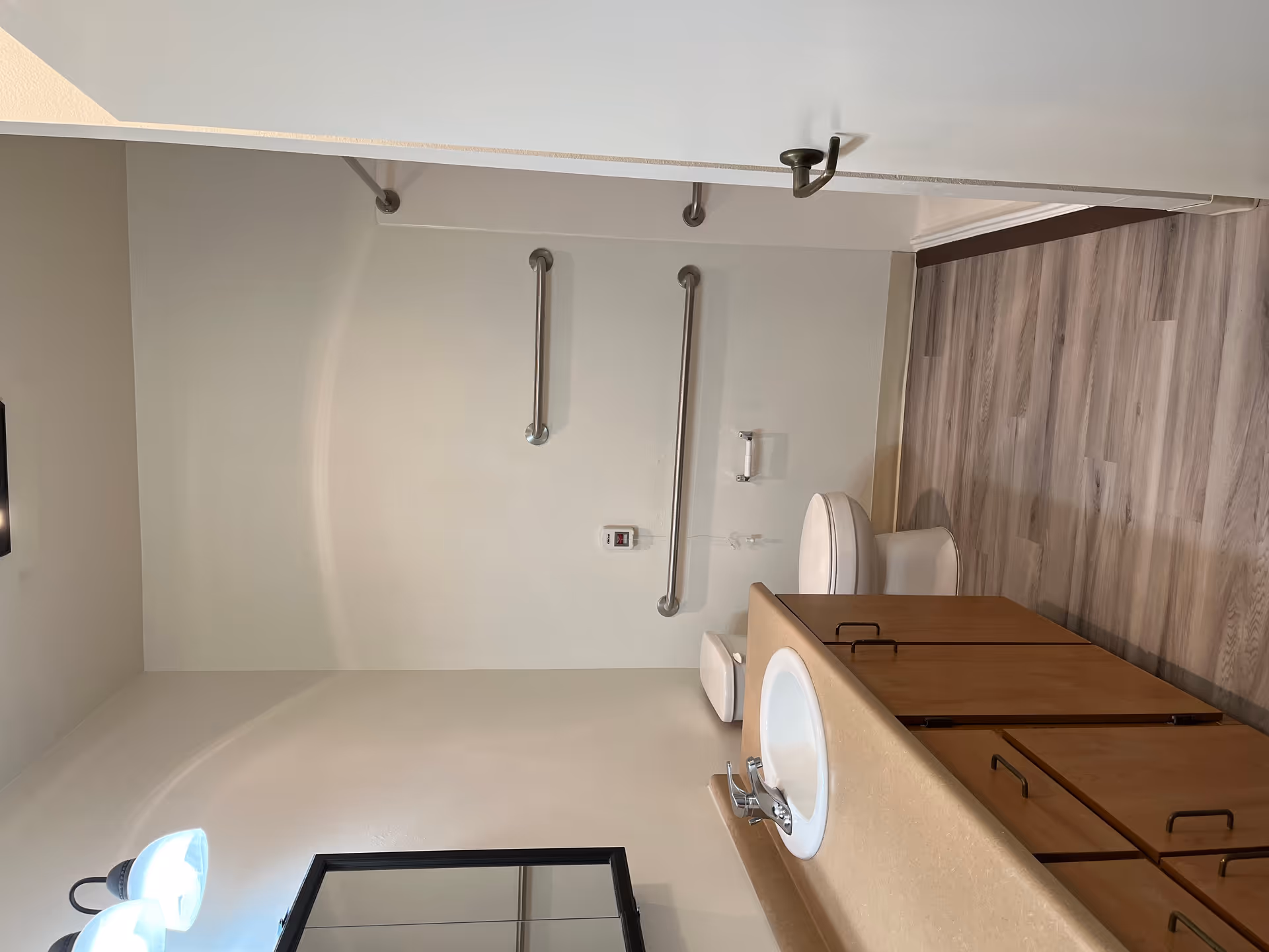 A clean bathroom with a wooden vanity featuring a white sink and faucet, a mirror with a black frame above the sink, a toilet with grab bars on the wall behind it, and a walk-in shower with a grab bar. The floor has light wood-patterned vinyl flooring, and the walls are painted white.