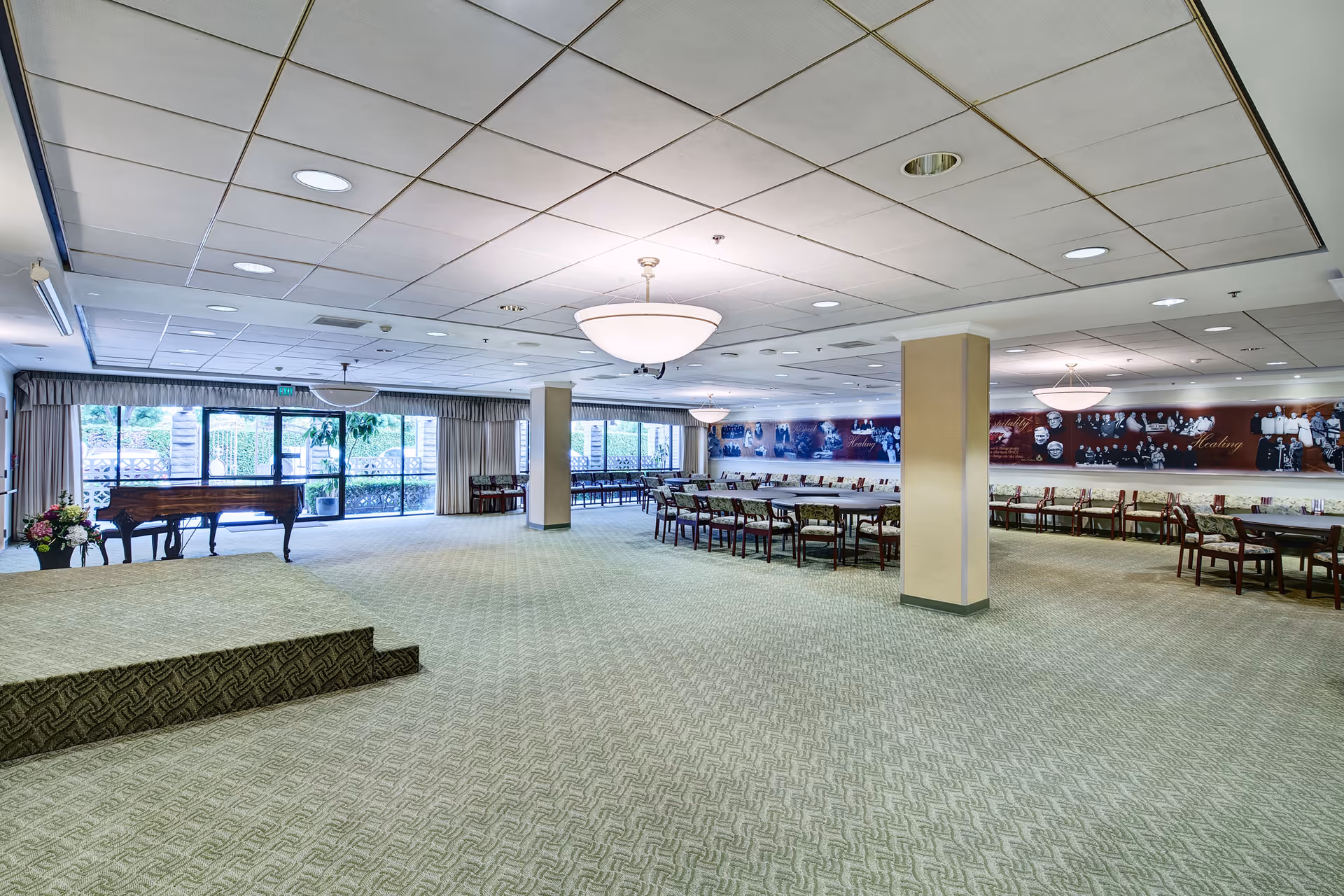 Large, carpeted room with a small stage, a grand piano, multiple tables and chairs arranged along the walls, large windows with curtains letting in natural light, and decorative wall art featuring historical photos and text.