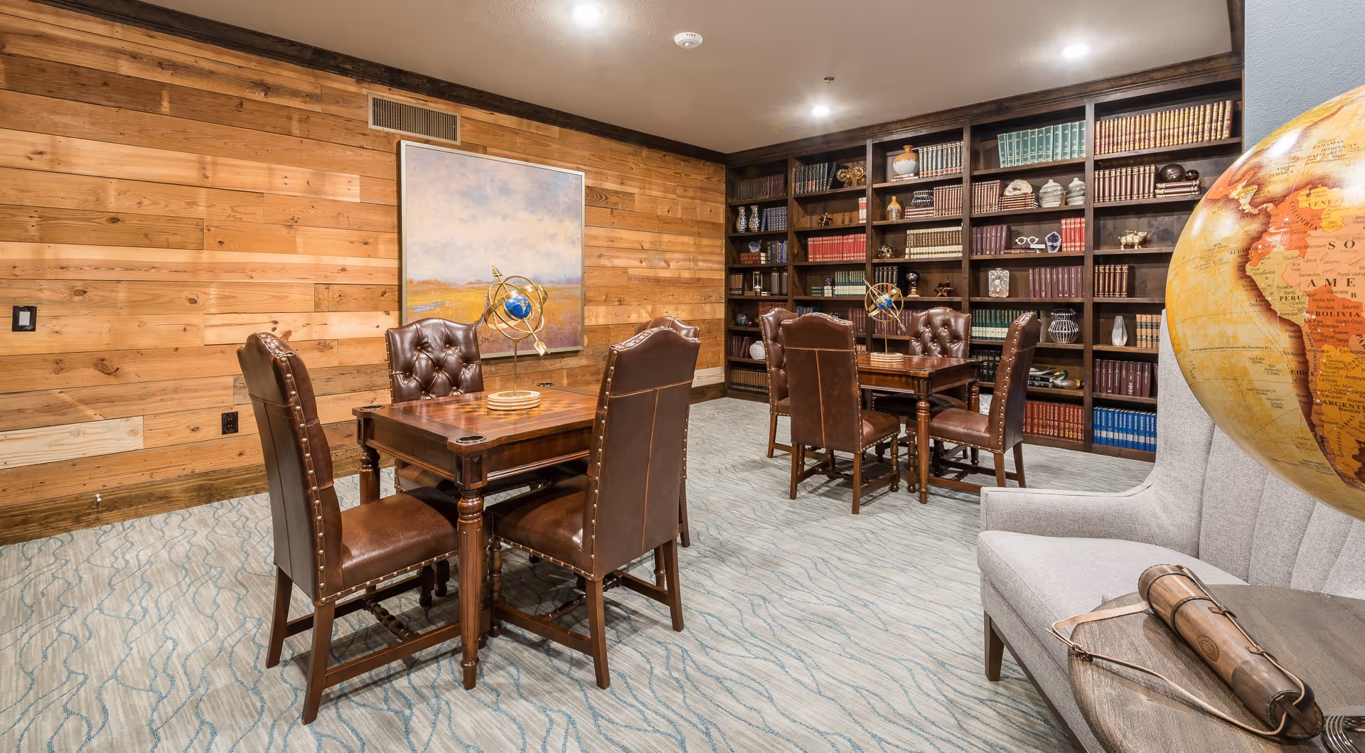 A cozy library or study room with wooden paneled walls and carpeted floor. The room features two wooden tables, each surrounded by four brown leather tufted chairs. A large bookshelf filled with books and decorative items covers the back wall. On the right side, there is a gray upholstered armchair next to a small round wooden table with a rolled-up scroll and a large globe partially visible.