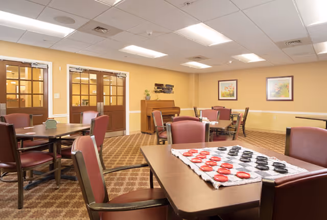 A well-lit common room with several tables and chairs arranged for social activities. One table in the foreground has a checkers game set up. The room features beige walls, carpeted flooring, framed floral artwork, and a piano against the far wall. Double wooden doors with glass panels are visible on the left side.