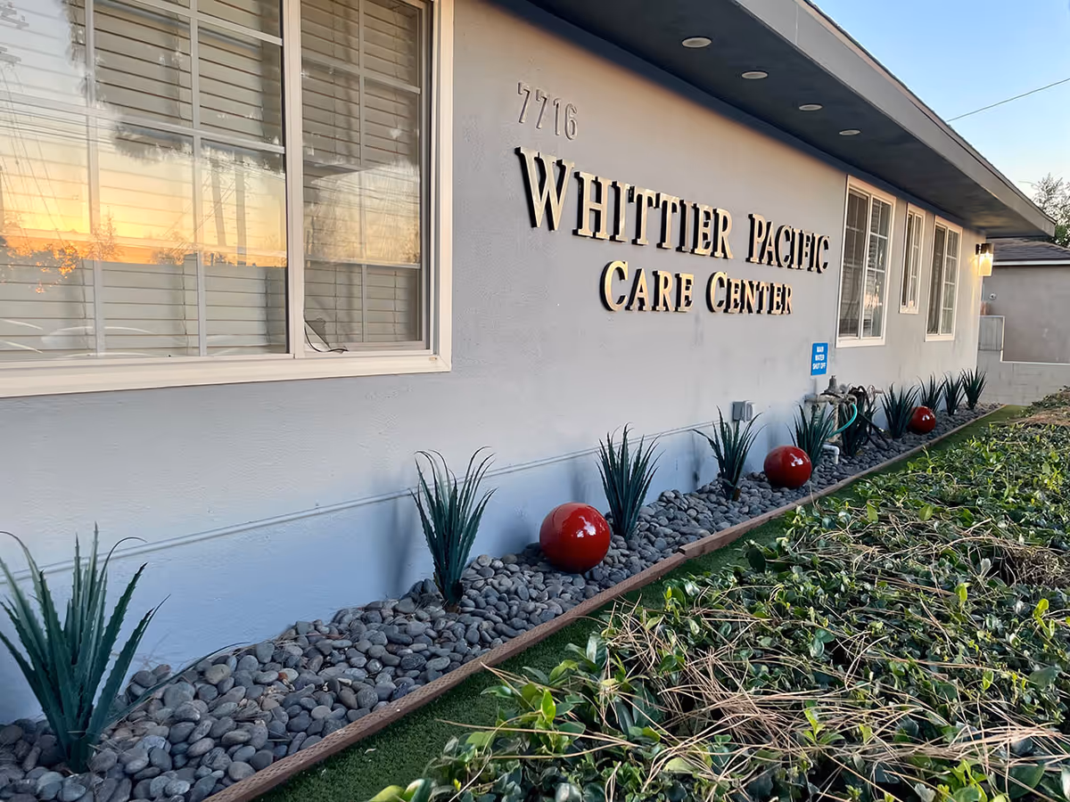 Exterior side view of the Whittier Pacific Care Center building with windows, a landscaped area featuring rocks, green plants, and red decorative spheres, under a clear sky at sunset.