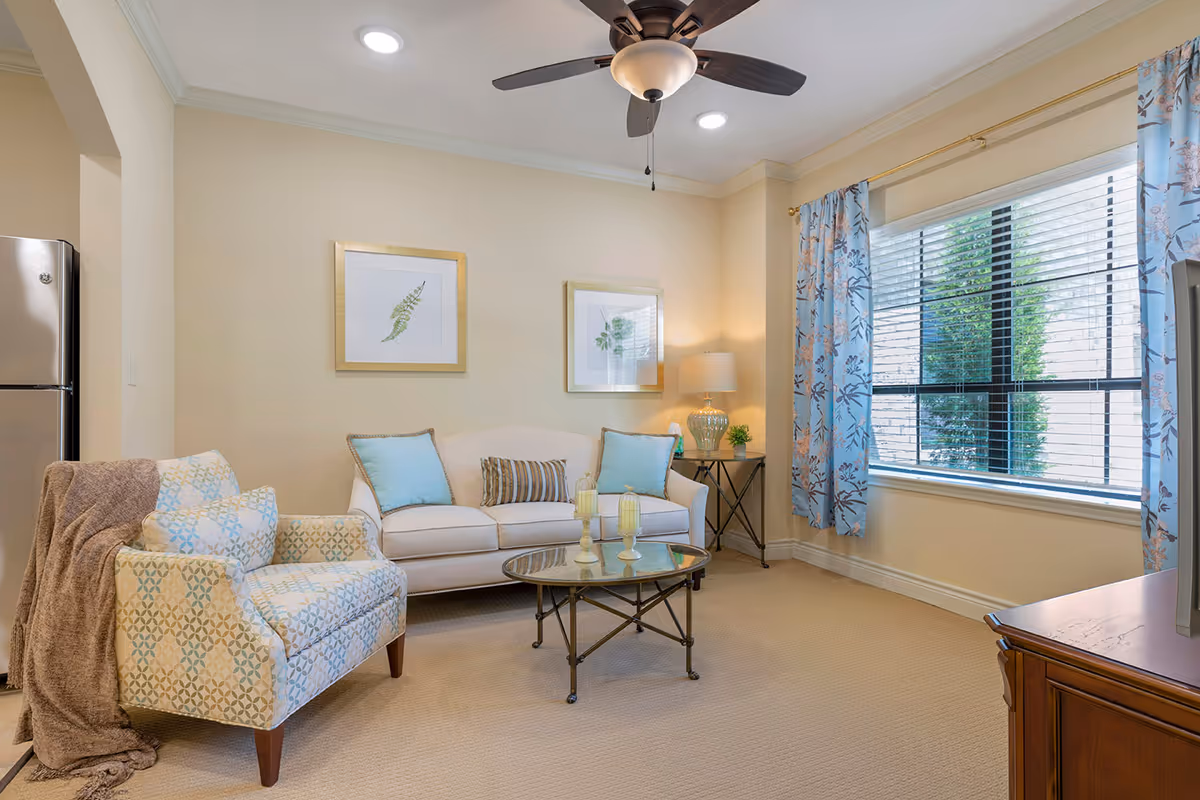 A cozy living room in a senior living facility featuring a beige sofa with blue and striped cushions, a patterned armchair with a brown throw blanket, a glass coffee table with decorative candles, a side table with a lamp and small plant, two framed botanical prints on the wall, a ceiling fan with lights, and a large window with blue floral curtains letting in natural light.