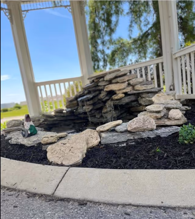 A small outdoor rock water feature with water gently flowing over stacked stones, surrounded by mulch and a concrete curb. The background shows a white wooden fence and green trees under a clear blue sky.