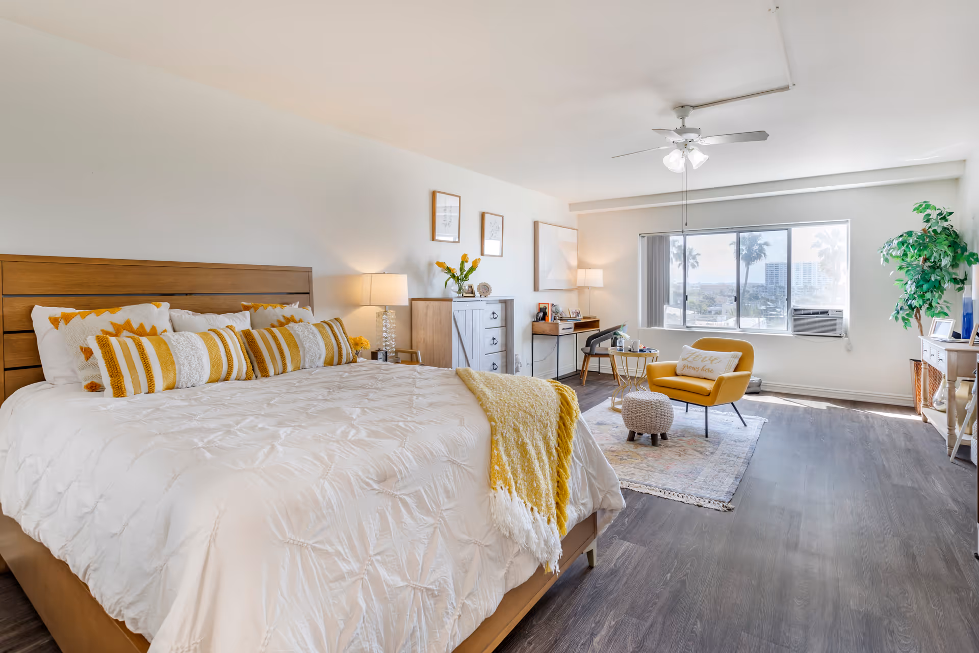 Bright and spacious bedroom with a large bed featuring white and yellow bedding and pillows. The room includes wooden furniture such as a dresser and desk, a yellow armchair with a small round table and ottoman on a rug near a large window with a view of palm trees and buildings. The room has wood flooring, a ceiling fan with lights, and a potted plant near the window.