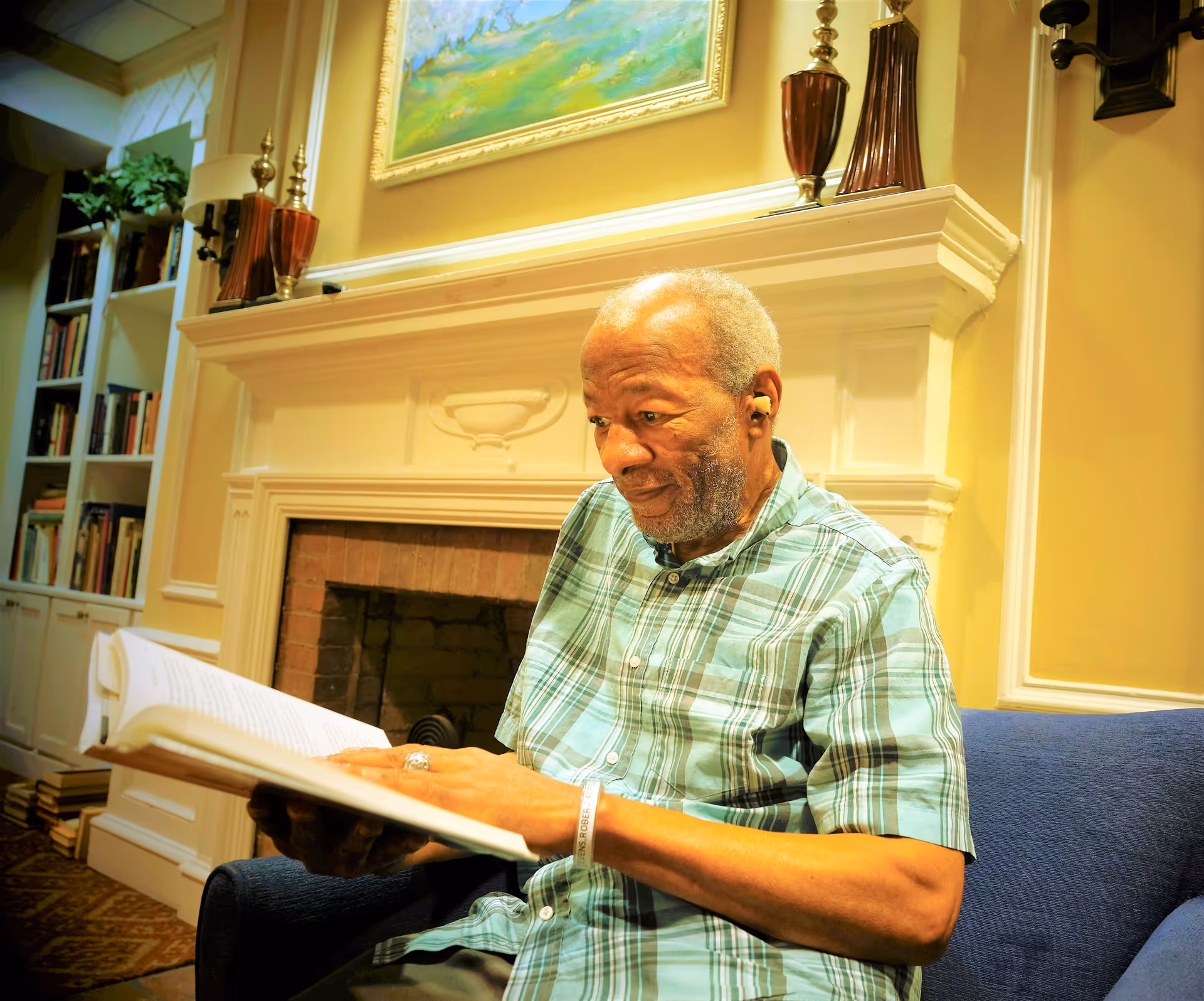 An elderly man wearing a green plaid shirt is sitting on a blue armchair reading a book in a cozy room with a white fireplace behind him. The room has warm yellow walls, decorative vases on the mantel, a framed painting above the fireplace, and a bookshelf filled with books to the left.