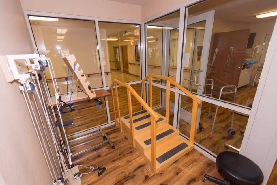A small indoor physical therapy area with wooden stairs equipped with handrails on both sides, exercise equipment including pulleys and resistance bands, and a black rolling stool. The room has wood-patterned flooring and large glass windows and doors looking into a hallway and another room with a walker visible.