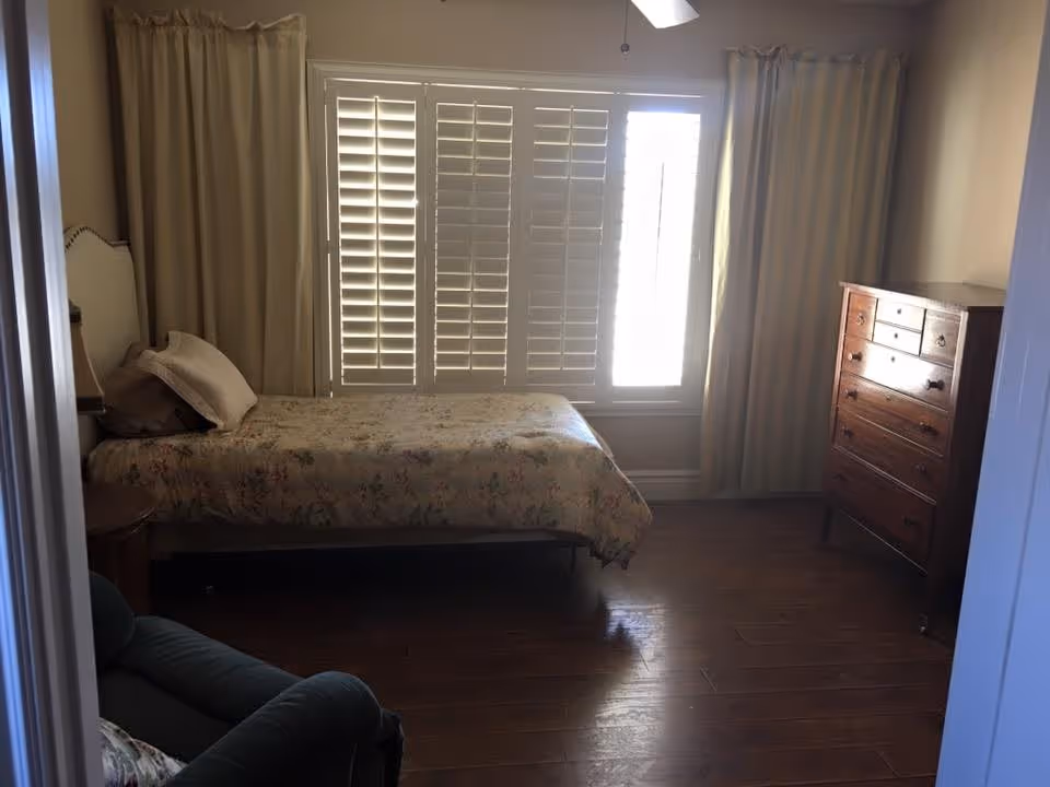 Sunlit bedroom with a single bed, floral bedspread, wooden dresser, shuttered window and a chair.
