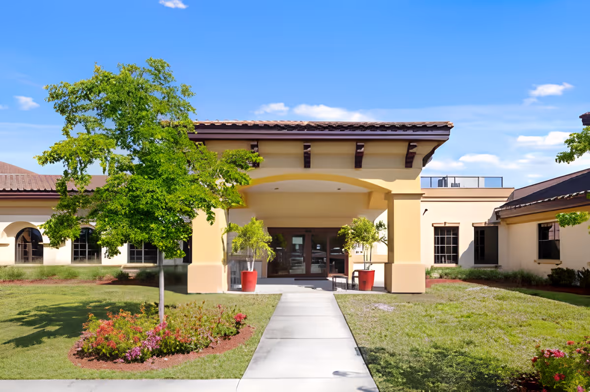 Front exterior view of a single-story building with a covered entrance supported by columns. There is a concrete walkway leading to the entrance, flanked by two potted plants with small trees. The building is surrounded by green grass, a tree, and flower beds under a clear blue sky.