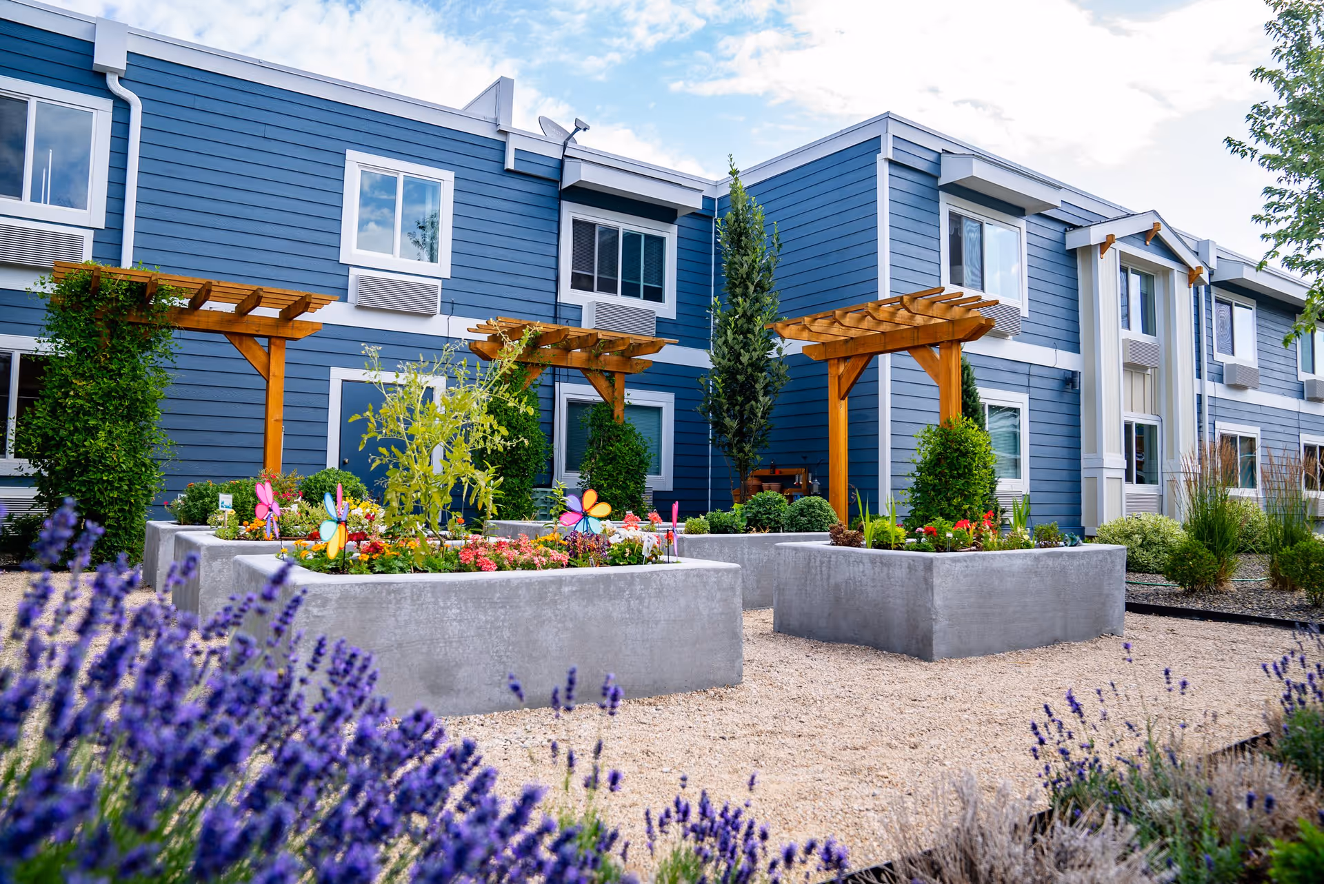 Outdoor garden area at Heatherwood Senior Living with raised concrete flower beds containing various plants and colorful pinwheels, wooden pergolas, and a blue two-story building in the background under a partly cloudy sky.