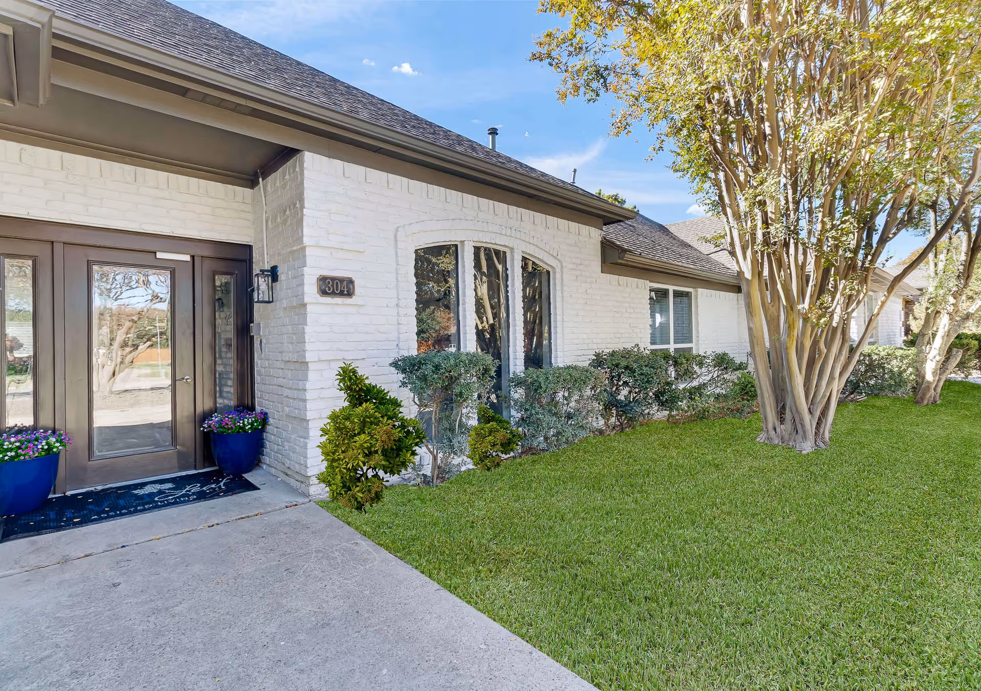Front entrance of a single-story white brick assisted living building with glass doors, an address plaque reading 304, potted flowers, and a manicured lawn with trees.