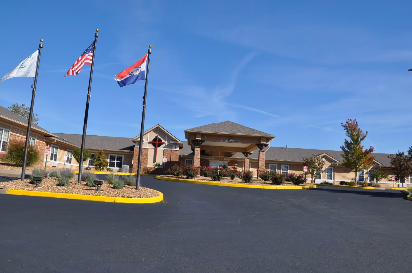 Exterior view of Arbor Hills Nursing and Rehabilitation Center building with a covered entrance, three flagpoles displaying flags, landscaped areas with shrubs and trees, and a clear blue sky.