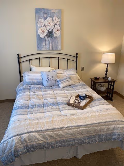 A neatly made bed with a metal headboard, striped bedding in shades of blue, beige, and white, and three pillows including one with the text 'live simply'. Above the bed hangs a painting of white flowers on a blue background. To the right of the bed is a wooden nightstand with a lamp, a small decorative item, and a tray with a cup and some papers on the bed.