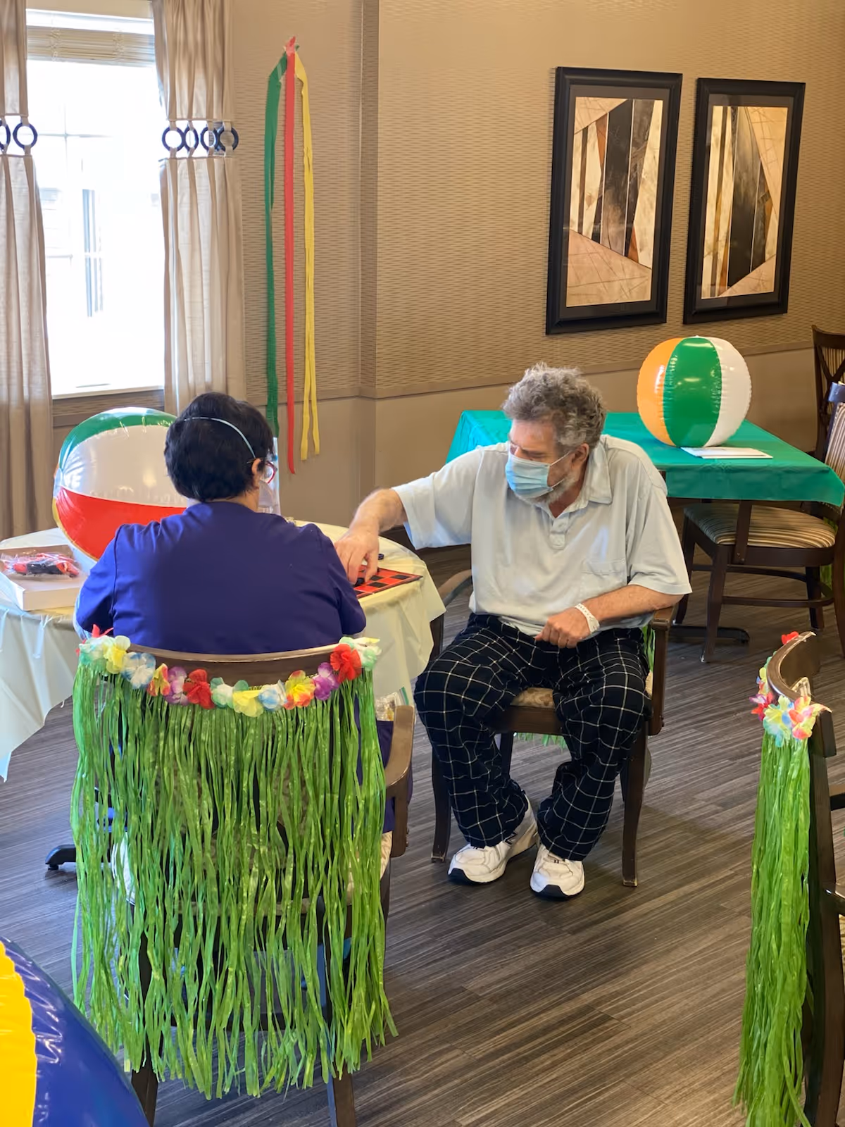 Two people sitting at a table in a decorated room, one wearing a face mask and the other with a green grass skirt and floral lei on the chair. They appear to be playing a board game. The room has beach balls, colorful streamers, and framed artwork on the wall.