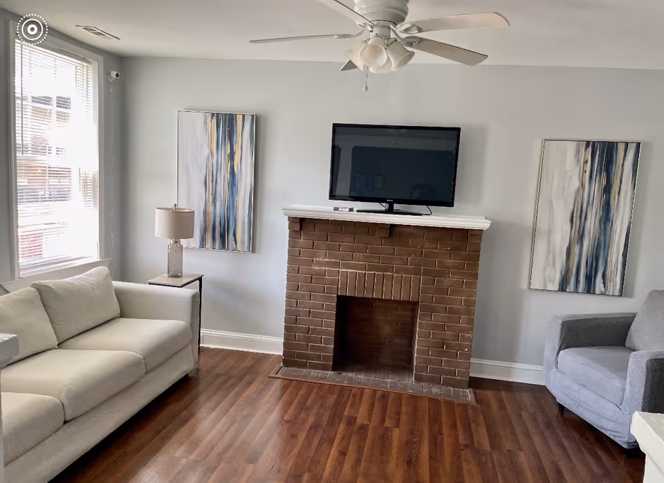 Bright living room with a white sofa, gray armchair, wall-mounted TV above a brick fireplace, wood floors, two abstract paintings and a ceiling fan.