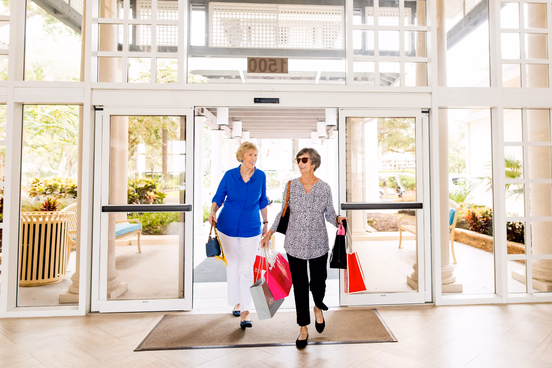 Two women carrying shopping bags walk through a glass automatic entrance into a bright lobby.