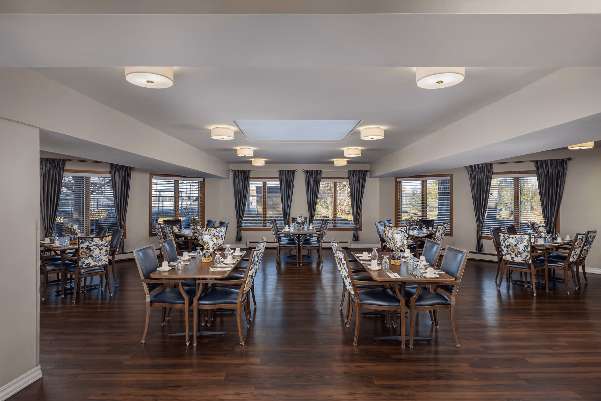 A spacious dining room with multiple wooden tables and chairs arranged neatly. Each table is set with white cups, saucers, and utensils. The room has large windows with dark curtains allowing natural light to enter. The floor is dark wood, and the ceiling has modern light fixtures and a skylight in the center.