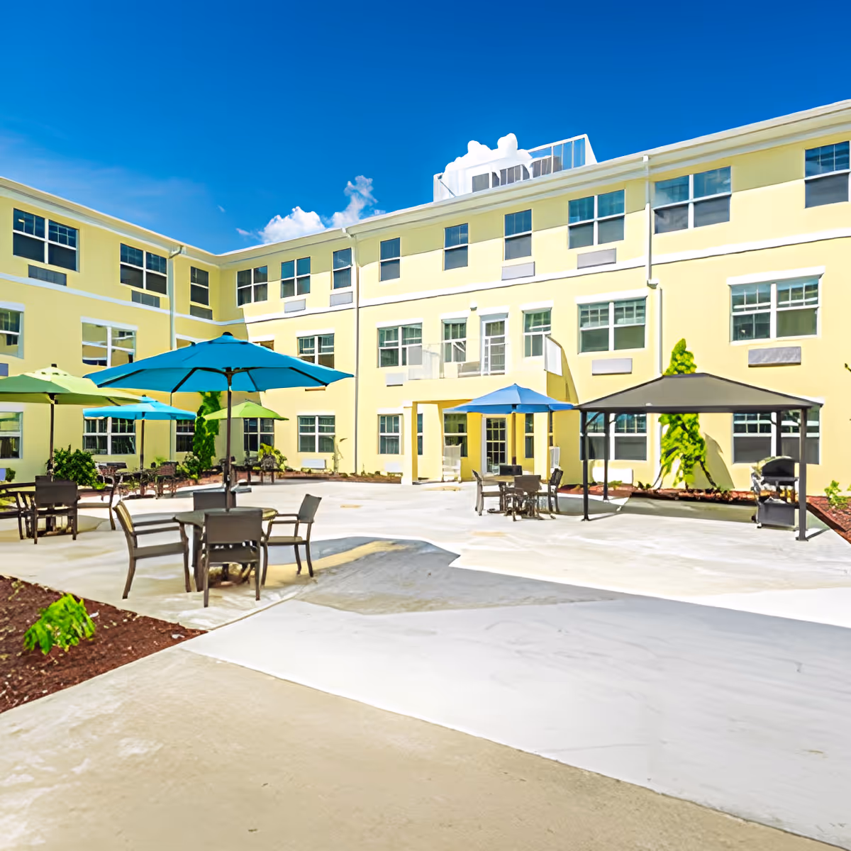 Outdoor courtyard area of a senior living community with multiple tables and chairs under colorful umbrellas, surrounded by a three-story yellow building under a clear blue sky.