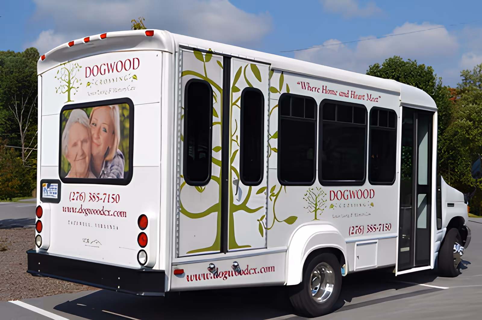 A white shuttle bus parked outdoors with the branding of Dogwood Crossing Senior Living And Memory Care. The bus features a large image of an elderly woman and a younger woman smiling together, along with green tree graphics and the facility's contact information and website.