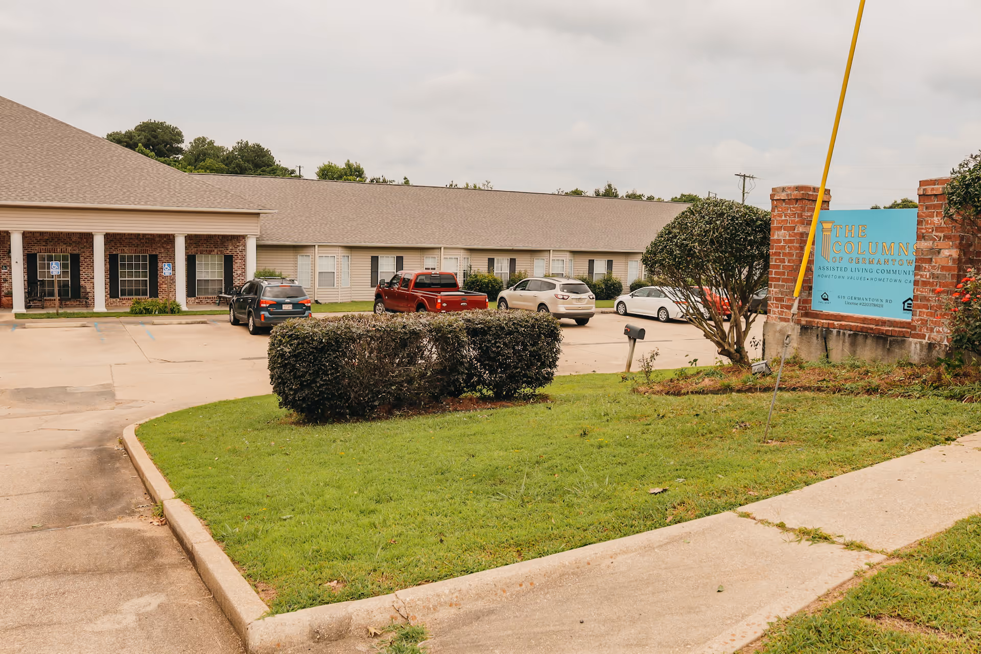 Front exterior of The Columns of Germantown assisted living building with parked cars, shrubbery, and a blue facility sign.