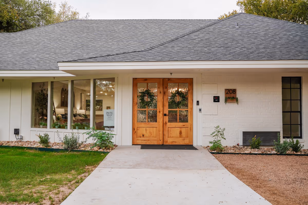 Front entrance of a single-story assisted living facility with double wooden doors, large windows, and the address number 208.