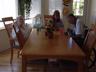 Three elderly people sitting around a wooden dining table in a room with large windows and wooden chairs. There is a vase with sunflowers on the table and some cups. The room has light-colored walls and wooden flooring.