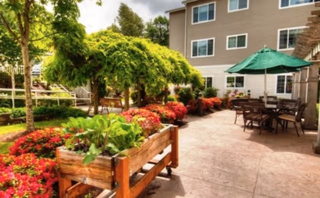 Outdoor patio area at Bay Pointe by Cogir featuring a paved walkway, wooden planter boxes with green plants, colorful flower beds, a large green umbrella shading a table with chairs, and a multi-story building in the background.
